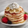 A tall stack of golden-brown banana protein pancakes on a white plate, topped with fresh banana slices, whipped cream, and served with a strawberry and a blackberry.