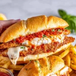 A hand holding a loaded chicken parmesan sandwich, with a crispy breaded chicken cutlet, chunky marinara sauce, and gooey melted mozzarella cheese on a hoagie roll, with french fries in the background.