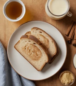 Slices of sourdough bread on a plate surrounded by ingredients like milk, cinnamon sticks, butter, and maple syrup.