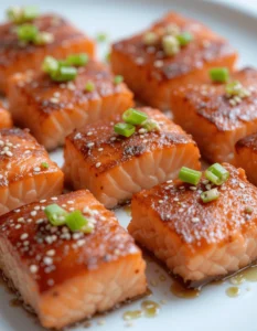 Multiple glazed salmon bites garnished with sesame seeds and green onions on a white plate.