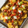 A close-up overhead shot of a platter of shredded beef nachos, generously topped with melted cheese, diced tomatoes, sliced jalapeños, avocado, and dollops of sour cream.