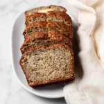 A loaf of moist banana bread made with a recipe using 2 bananas, sliced on a white oval plate next to a cream-colored napkin