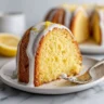 A close-up slice of moist lemon cream cheese pound cake on a white plate, topped with a thick white lemon glaze and fresh lemon zest, with the rest of the Bundt cake in the background