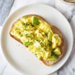 An overhead shot of a single slice of toast on a white plate, piled high with fluffy avocado scrambled eggs and garnished with fresh herbs