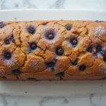 Whole blueberry loaf cake with a golden crust and sugar topping on a white platter.