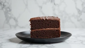 A slice of moist chocolate cake with creamy chocolate frosting on a black plate, placed on a white marble background.