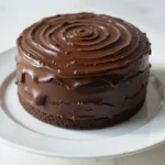 A round, moist chocolate cake covered in glossy chocolate ganache with a swirl pattern, served on a white plate over a marble surface.