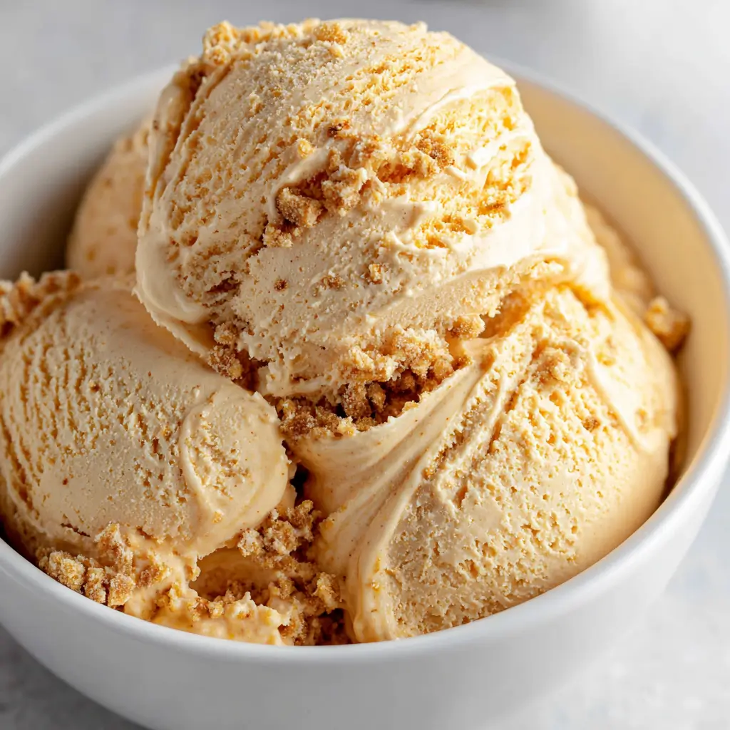 Three scoops of creamy pumpkin pie ice cream in a white bowl, generously sprinkled with crumbled pie crust.