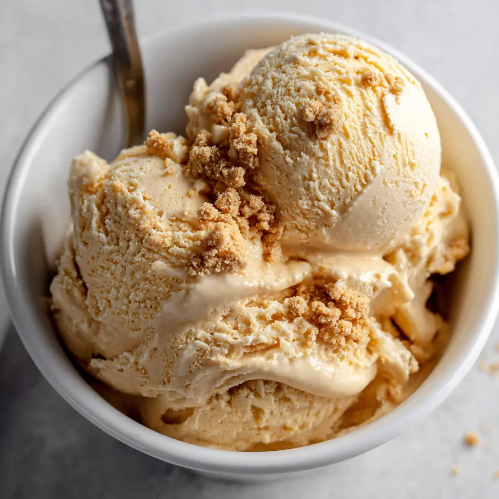 Top-down view of a white bowl filled with three scoops of creamy pumpkin pie ice cream, garnished with golden-brown pie crust crumbles and a silver spoon.