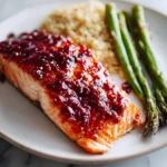 A perfectly cooked fillet of raspberry glazed salmon on a white plate, served with fluffy quinoa and roasted asparagus spears.