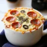 Close-up of a pizza in a mug with melted cheese, pepperoni, olives, and herbs