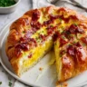 A golden-baked croissant breakfast pie on a white plate, generously filled with fluffy scrambled eggs and topped with crispy bacon and fresh chives, with one slice cut out.