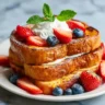A tall stack of golden-brown eggless French toast on a white plate, topped with whipped cream, fresh sliced strawberries, blueberries, and a mint sprig