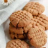 A stack of keto almond cookies with a classic criss-cross pattern, next to a light blue cloth and white flowers, suggesting a fresh and delightful treat.