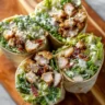 Four crispy chicken Caesar wraps, standing upright on a wooden cutting board, each revealing their delicious filling of golden-brown fried chicken, crisp green lettuce, creamy Caesar dressing, and bacon bits, topped with grated cheese.