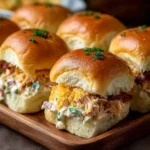 A wooden tray holds several golden-baked crack chicken sliders, generously filled with a creamy chicken mixture, crispy bacon, and topped with a sprinkle of seasoning and fresh green herbs.