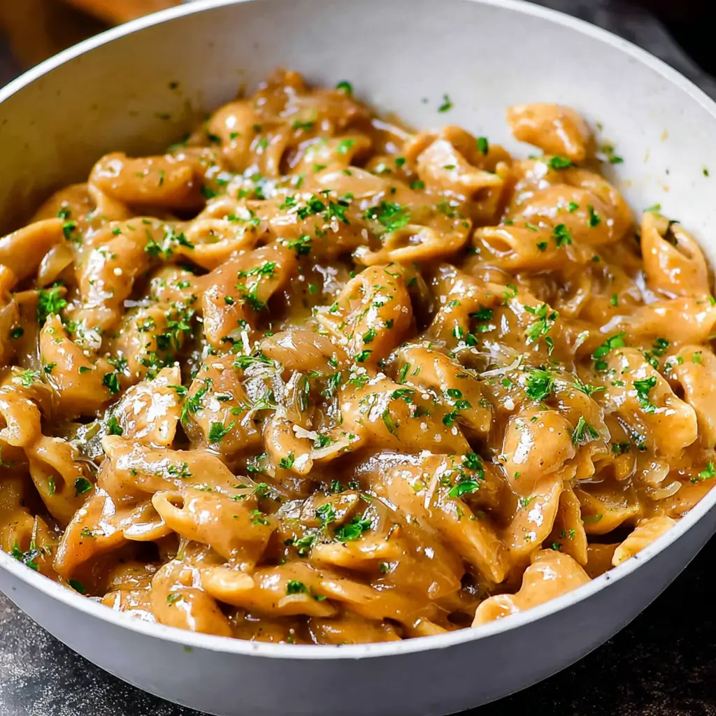 A close-up shot of a bowl filled with creamy one pot French onion pasta, featuring shell pasta coated in a rich, savory sauce, garnished with finely chopped fresh parsley and grated cheese.