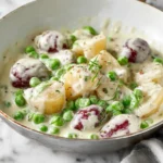 A close-up of a skillet filled with creamy peas and potatoes, featuring tender new red and white potatoes and bright green peas in a savory white sauce, garnished with fresh herbs