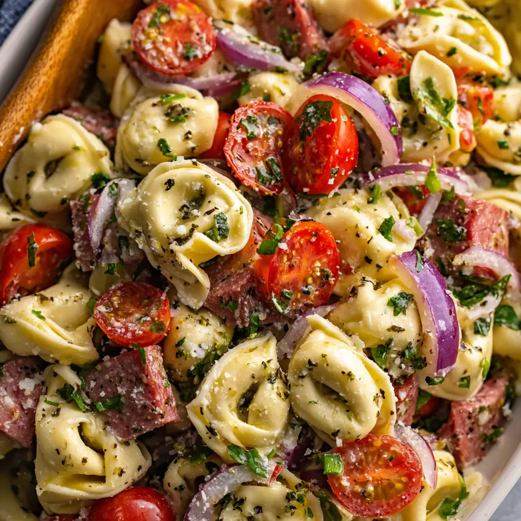 A close-up macro shot of a tortellini pasta salad with Italian dressing, highlighting the cheese tortellini, salami, fresh cherry tomatoes, and red onion.