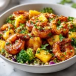 A close-up shot of a healthy Caribbean shrimp bowl in a white bowl, filled with seasoned shrimp, pineapple chunks, and broccoli florets over couscous.