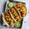 An overhead view of three golden-brown coconut shrimp tacos on a metal tray, generously topped with fresh mango salsa, a creamy orange sauce, and cilantro, with lime wedges on the side