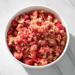 A white bowl filled with homemade strawberry crunch topping, featuring vibrant red strawberry pieces and golden cookie crumbles, set against a white marble background