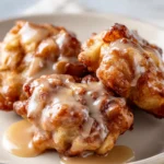 A close-up shot of three golden-brown, crispy air fryer apple fritters on a cream-white plate, generously drizzled with a sweet vanilla glaze.
