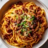 A close-up overhead shot of Cowboy Spaghetti in a white bowl, featuring spaghetti noodles tossed in a hearty meat sauce and garnished with fresh green onions and grated Parmesan cheese.