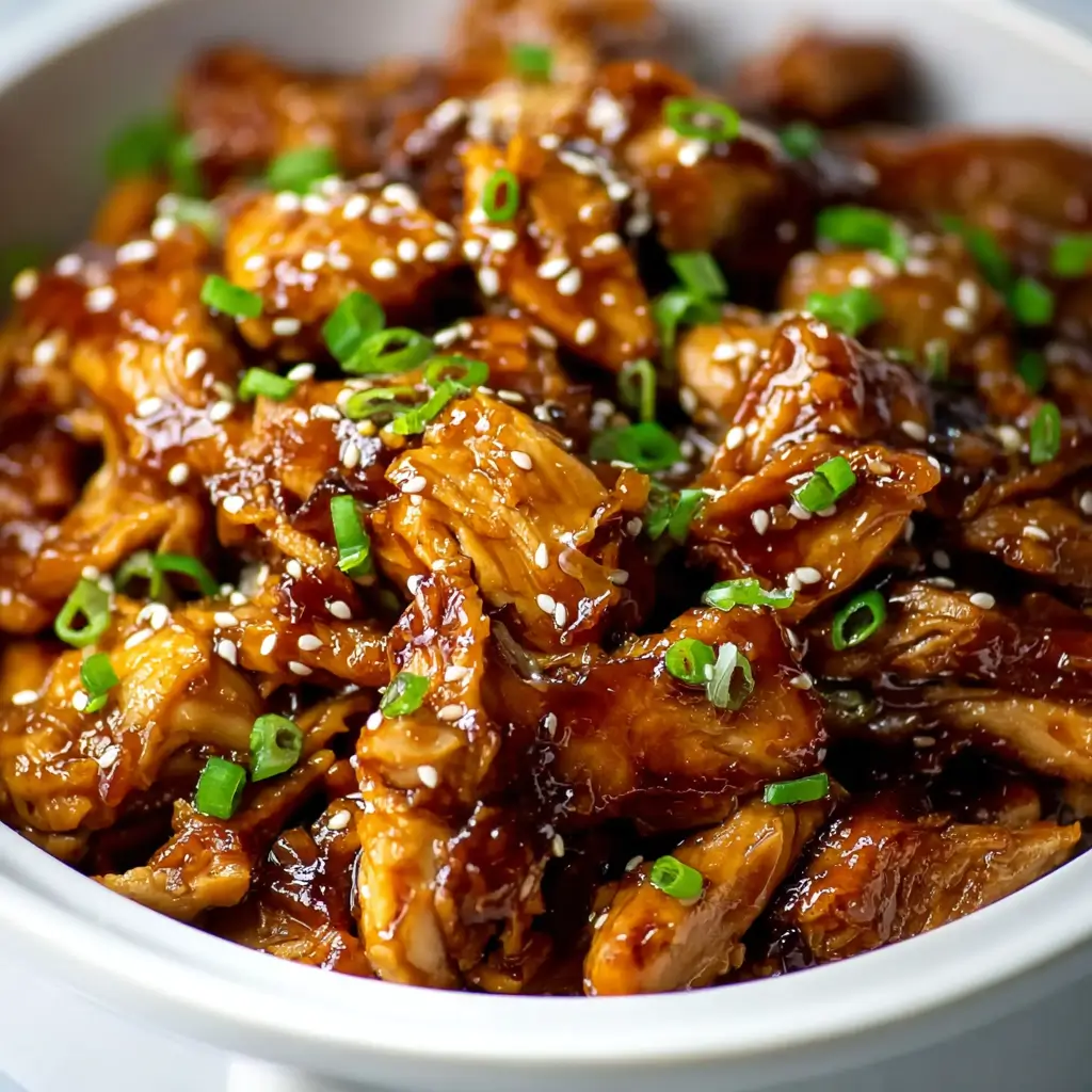 A close-up shot of a white bowl filled with shredded Crockpot Teriyaki Chicken, garnished with sesame seeds and sliced green onions.