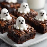 A close-up of a rich, fudgy ghost brownie on a white plate, topped with a swirl of chocolate ganache and a cute marshmallow ghost with black candy eyes.