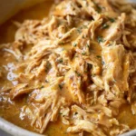 A close-up bowl of tender, shredded gluten-free crockpot chicken smothered in a thick, savory gravy and garnished with fresh parsley.