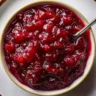 An overhead shot of a white bowl filled with vibrant, chunky Homemade Cranberry Orange Sauce, with a spoon resting inside.