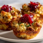 A close-up of three golden-brown leftover stuffing muffins on a white plate, each topped with a dollop of cranberry sauce and garnished with fresh herbs.