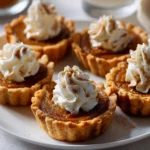 A close-up of five individual mini pumpkin pies on a white plate, each topped with a swirl of whipped cream and a sprinkle of cinnamon.