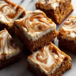 A close-up of several moist pumpkin bars with a thick layer of cream cheese frosting and a caramel swirl, stacked on a white marble surface.