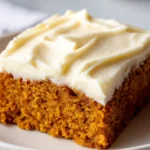 A close-up shot of a single square slice of a moist pumpkin bar on a cream-white plate, topped with a thick, generous layer of swirled cream cheese frosting.