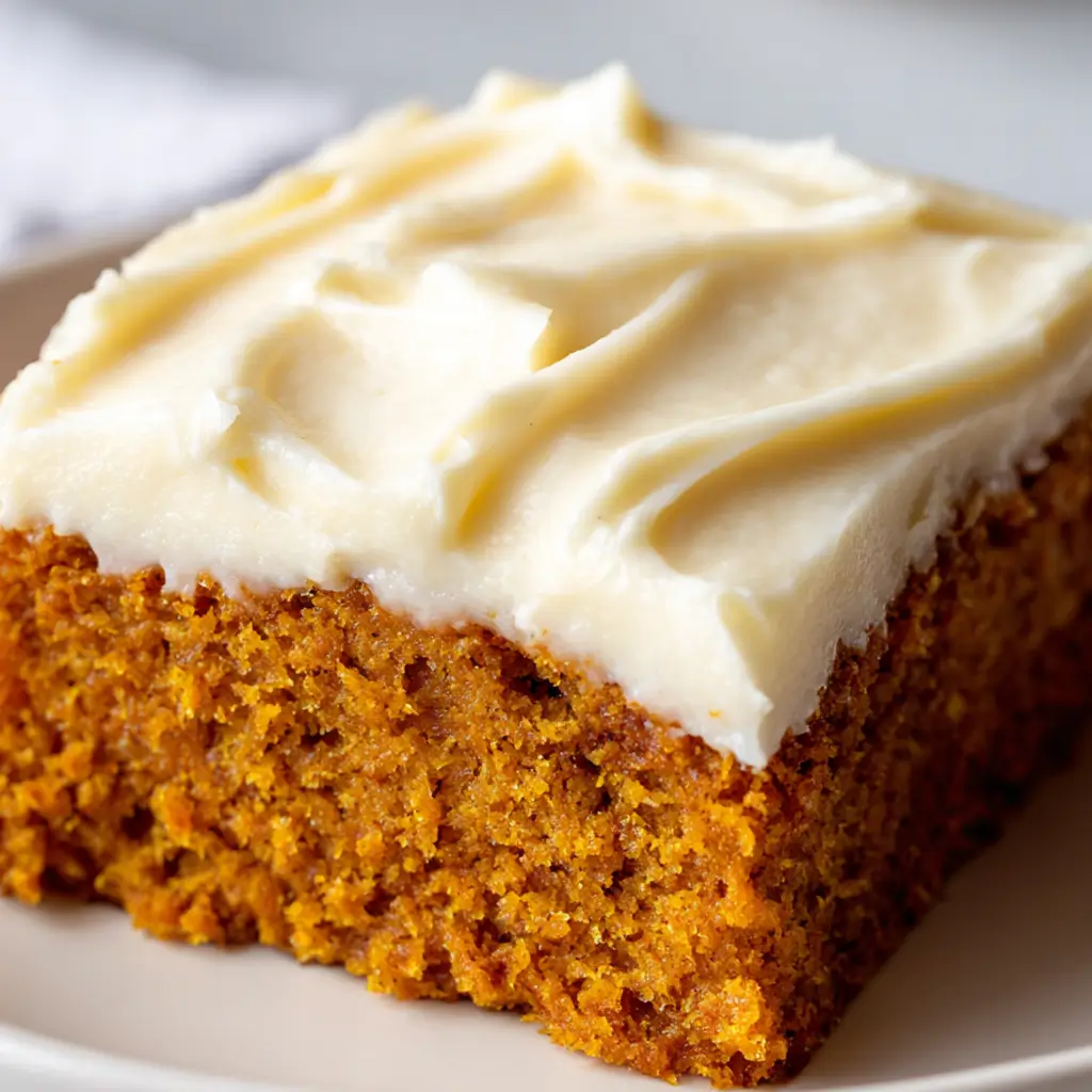A close-up shot of a single square slice of a moist pumpkin bar on a cream-white plate, topped with a thick, generous layer of swirled cream cheese frosting.