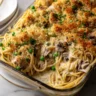 A close-up shot of a glass baking dish filled with creamy Turkey Tetrazzini, showing spaghetti, chunks of turkey, peas, and mushrooms in a cream sauce, topped with golden-brown breadcrumbs and fresh parsley.