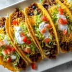 Four crispy ground beef tacos lined up on a white platter, each filled with seasoned beef, shredded lettuce, cheddar cheese, sour cream, and diced tomatoes.