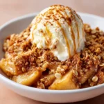 A close-up photo of apple crisp with oats, showing baked spiced apples topped with a golden oat crumble, finished with a scoop of vanilla ice cream drizzled with caramel sauce.