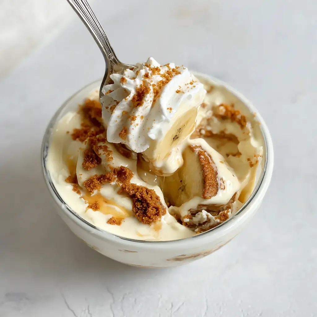 Small glass bowl of Biscoff banana pudding with layers of creamy custard, banana slices, whipped topping, and crumbled cookies, with a spoon lifting a bite above the bowl.