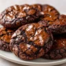 Chewy, crackly brownie cookies stacked on a white plate, with signature shiny tops and rich dark chocolate color.