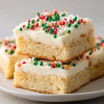 Frosted Christmas sugar cookie bars cut into squares, topped with creamy white icing and festive red, green, and white holiday sprinkles, stacked on a white plate.