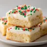 Frosted Christmas sugar cookie bars cut into squares, topped with creamy white icing and festive red, green, and white holiday sprinkles, stacked on a white plate.