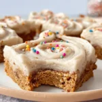 Cinnamon Roll Bars on a plate, featuring a moist cinnamon cookie base, a generous swirl of creamy frosting, and colorful rainbow sprinkles on top. One bar in the foreground has a bite taken out, showing its soft texture.