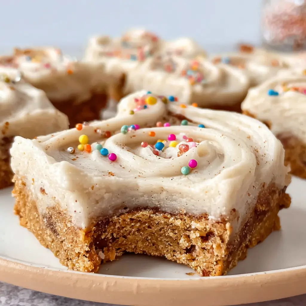 Cinnamon Roll Bars on a plate, featuring a moist cinnamon cookie base, a generous swirl of creamy frosting, and colorful rainbow sprinkles on top. One bar in the foreground has a bite taken out, showing its soft texture.