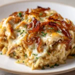 A rich, golden-brown serving of French Onion Chicken Orzo Casserole on a white plate, featuring tender orzo and shredded chicken enveloped in creamy cheese sauce, topped with a generous layer of caramelized onions and a sprinkle of fresh herbs.