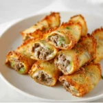 A plate of crispy, golden-brown Keto Philly cheesesteak roll-ups, cut in half to show the savory ground beef, melted cheese, and bits of fresh green pepper inside.