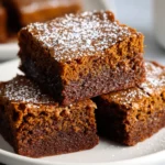 Square gingerbread brownies on a plate, beautifully dense with a deep brown color, soft and fudgy interior, topped with a dusting of powdered sugar for the holidays.