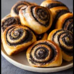 Poppyseed buns arranged on a ceramic plate, featuring golden-brown pastry swirled with a generous layer of glossy black poppyseed filling.